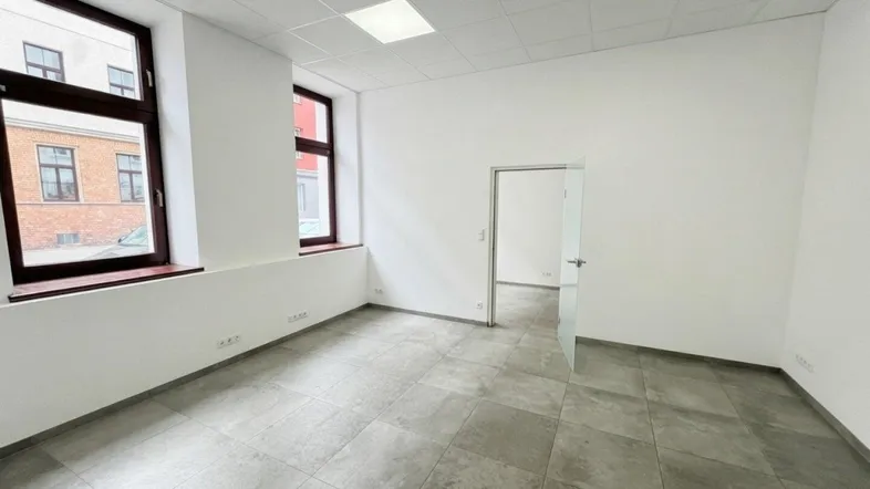 Heller Büroraum mit großen Fenstern, grauen Fliesen und einer Glastür zu einem weiteren Bereich.