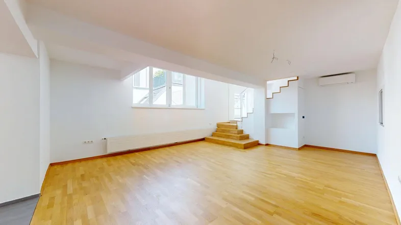 Geräumiger Wohnbereich mit hellem Holzfußboden, großen Fenstern und einer Treppe zum oberen Stockwerk.