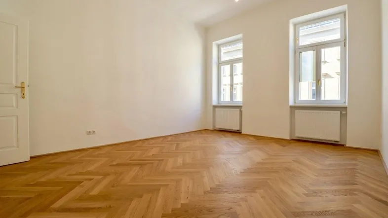 Helles Zimmer mit Fischgrätparkettboden, weißen Wänden und zwei Fenstern, bereit zum Einzug.