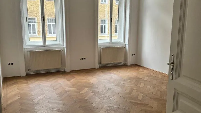 Helles Zimmer mit Fischgrätparkettboden und zwei hohen Fenstern, die viel Tageslicht hereinlassen.