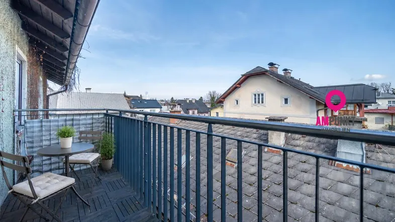Gemütlicher Balkon mit Sitzgelegenheiten und weitem Blick über die Dächer der umliegenden Häuser unter blauem Himmel.
