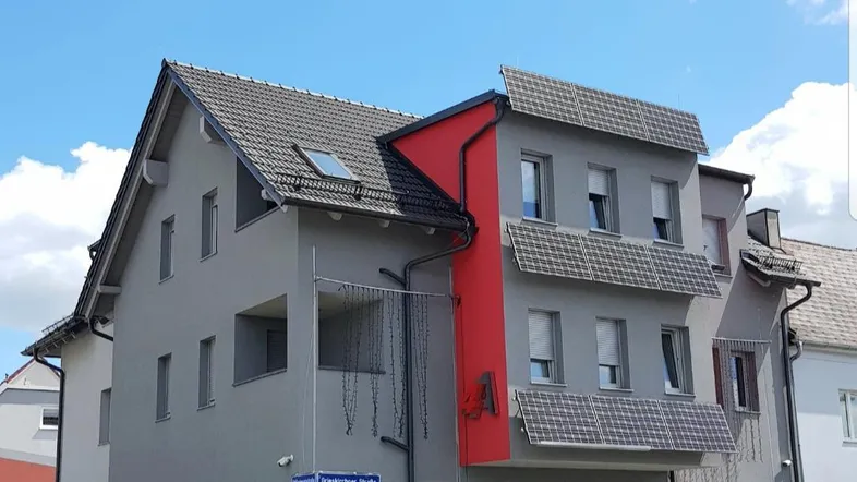 Modernes Mehrfamilienhaus mit grauer Fassade, roten Akzenten und integrierten Solarpaneelen auf dem Dach.