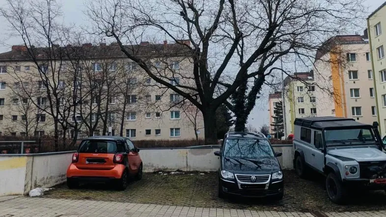 Außenansicht eines Parkplatzes mit mehreren Autos vor Wohngebäuden unter einem kahlen Baum.