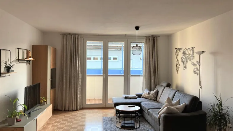 Helles Wohnzimmer mit Parkettboden, bequemer Couch und Zugang zum Balkon.