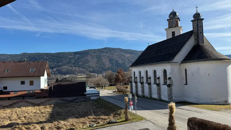 Blick auf eine Kirche mit Bergen im Hintergrund und umliegenden Häusern an einem sonnigen Tag.