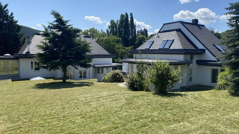 Zwei moderne Häuser mit dunklen Dächern und weißer Fassade, umgeben von einem weitläufigen Rasen und Bäumen.