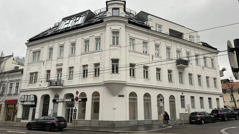 Weißes Eckgebäude mit klassischer Architektur und modernen Dachaufbauten an einer Stadtstraße.