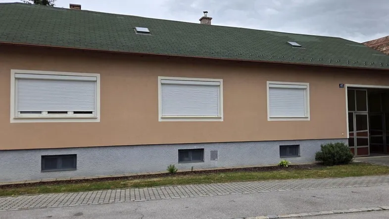 Vorderansicht des Hauses mit beiger Fassade, grünem Dach und geschlossenen Rollläden an den Fenstern.