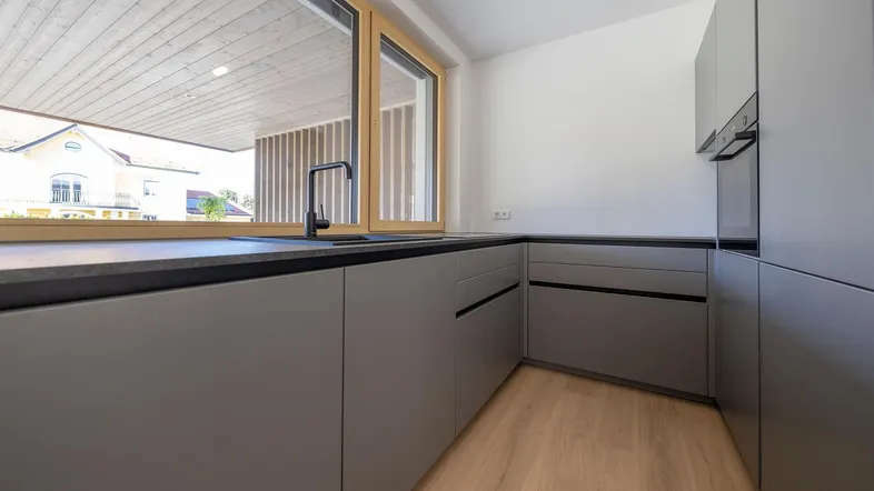Moderne Küche mit grauen Schränken, dunkler Arbeitsplatte und großem Fenster.
