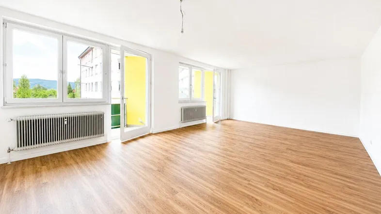 Helles Wohnzimmer mit Laminatboden, großen Fenstern und Zugang zu einem Balkon.