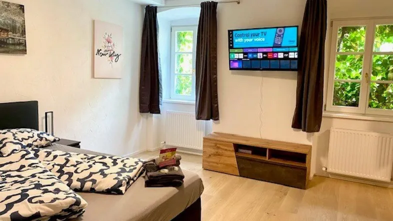 Gemütliches Schlafzimmer mit Holzboden, Doppelbett, Fernseher und Blick ins Grüne durch die Fenster.