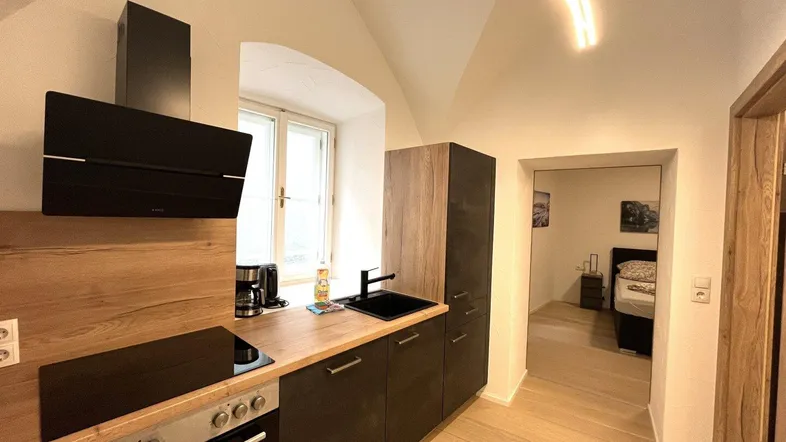 Moderne Küche mit Holzarbeitsplatte, dunklen Schränken und Blick in ein Schlafzimmer.