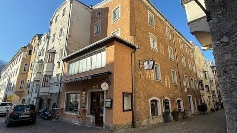 Historisches Gebäude an einer gepflasterten Straße mit Geschäften im Erdgeschoss und Wohnungen darüber, unter blauem Himmel.