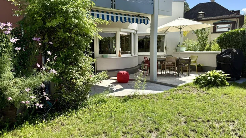 Große Terrasse mit Gartenmöbeln, Sonnenschirm und Grillbereich, umgeben von Grünflächen.