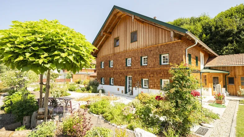 Traditionelles Holzhaus mit gepflegtem Garten und Sitzbereich unter einem Baum an einem sonnigen Tag.