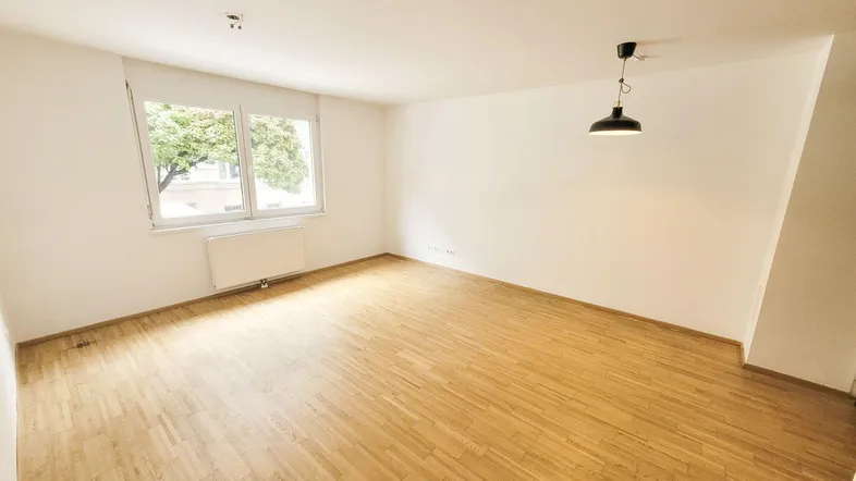 Helles, leeres Zimmer mit Parkettboden und einem Fenster, das viel Tageslicht hereinlässt.