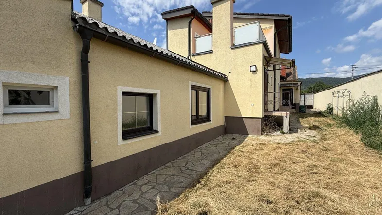 Seitenansicht des Hauses mit gelber Fassade und einem ungenutzten Gartenbereich unter blauem Himmel.