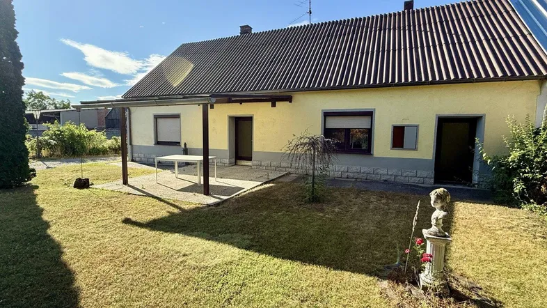 Hausrückseite mit großem Garten, überdachter Terrasse und Sitzbereich unter blauem Himmel.