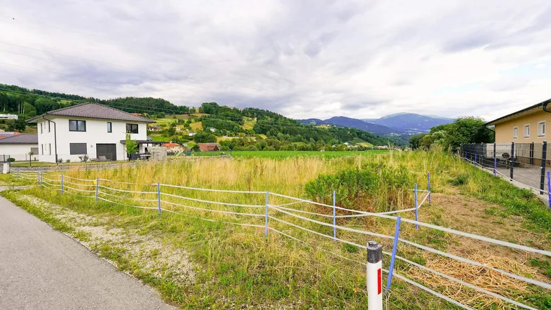 Blick auf ein unbebautes Grundstück mit umliegenden Häusern und einer grünen Hügellandschaft im Hintergrund.