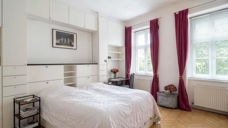 Helles Schlafzimmer mit Einbauschränken, Parkettboden und großen Fenstern mit roten Vorhängen.