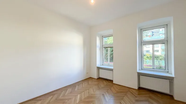 Helles Zimmer mit Fischgrätparkett und zwei Fenstern, ideal als Wohn- oder Schlafzimmer.