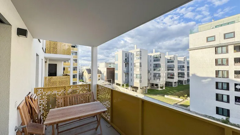 Möblierter Balkon mit Holztisch und Stühlen, bietet Blick auf moderne Wohngebäude und Grünflächen.