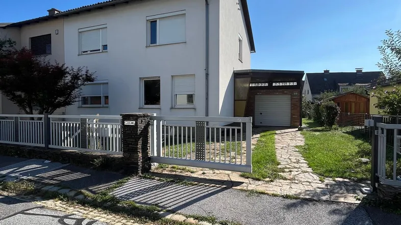Weißes Einfamilienhaus mit Vorgarten, Zaun und Einfahrt unter blauem Himmel.