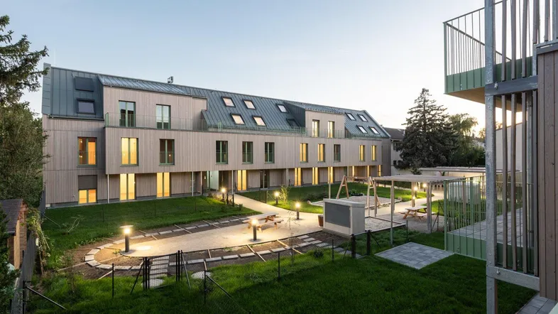 Moderne Wohnanlage mit Holzfassade, Balkonen und beleuchtetem Gemeinschaftsgarten am Abend.