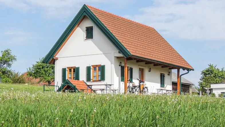 Kleines weißes Haus mit rotem Dach und grünen Fensterläden, umgeben von einer blühenden Wiese unter blauem Himmel.