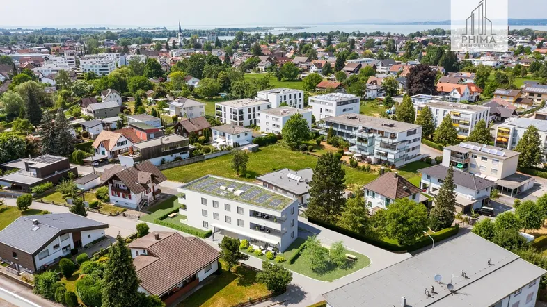 Luftaufnahme einer modernen Wohnanlage in grüner Umgebung mit See im Hintergrund.