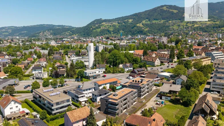 Luftaufnahme einer Stadt mit Wohngebäuden, Straßen und grünen Hügeln im Hintergrund unter blauem Himmel.