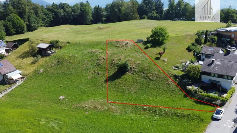 Luftaufnahme eines Baugrundstücks mit roter Umrandung, umgeben von grünen Hügeln und Häusern.