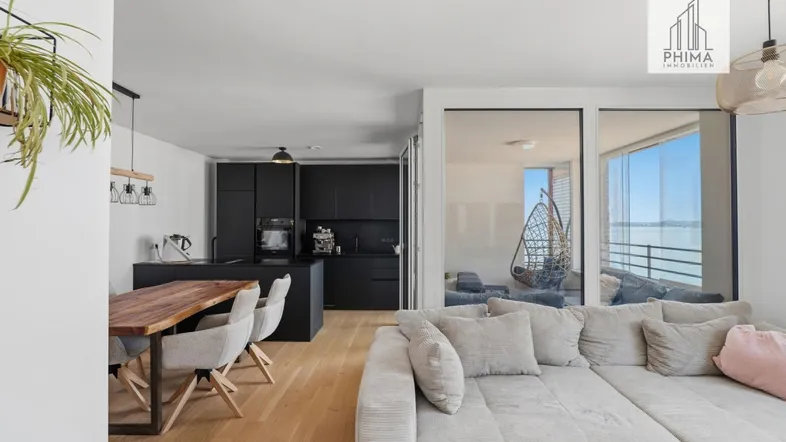 Moderner offener Wohn-Essbereich mit schwarzer Küche, Holzboden und Zugang zum verglasten Balkon.