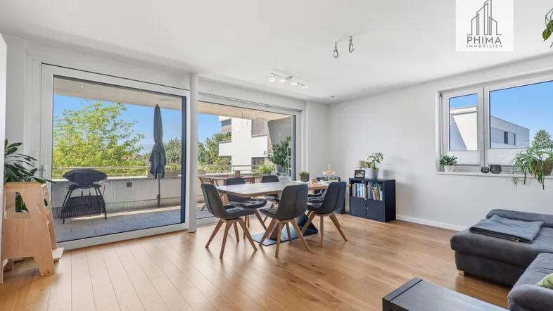 Helles Wohn- und Esszimmer mit Holzboden, Zugang zum Balkon und Blick ins Grüne.