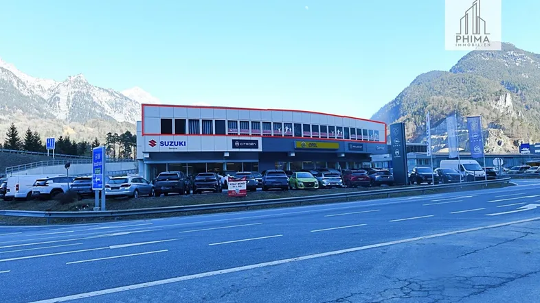 Außenansicht eines Autohauses mit Parkplätzen und Bergen im Hintergrund unter blauem Himmel.