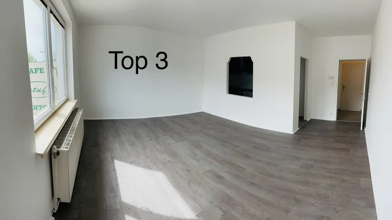 Helles, leeres Wohnzimmer mit modernem Laminatboden und großem Fenster.