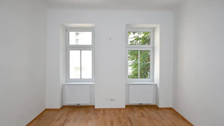 Helles Zimmer mit zwei Fenstern, Parkettboden und weißen Wänden, bereit zur individuellen Gestaltung.