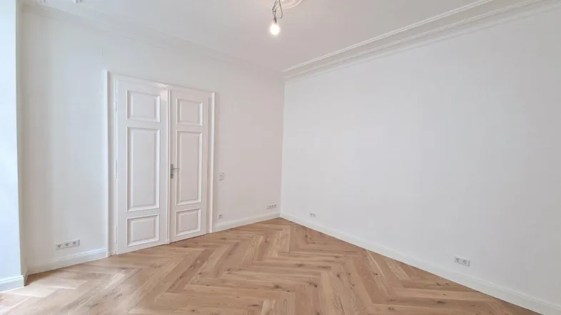 Helles Zimmer mit Fischgrätparkett, hohen Decken und eleganten Doppeltüren, ideal für Wohn- oder Schlafbereich.