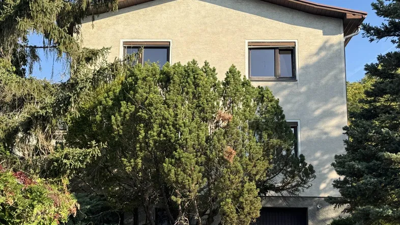 Hausfassade mit Bäumen und Sträuchern im Vordergrund, Garage im Erdgeschoss und Rollläden an den Fenstern.