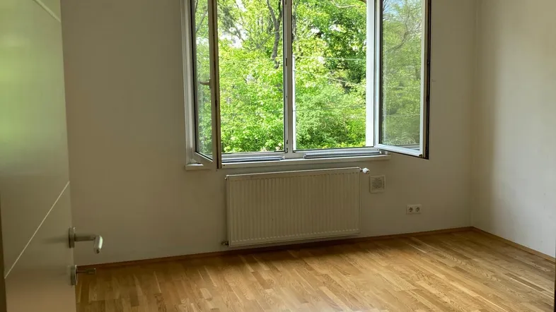 Helles Zimmer mit Parkettboden und Blick ins Grüne durch das geöffnete Fenster.