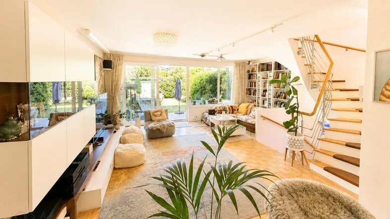 Helles Wohnzimmer mit Parkettboden, großen Fenstern zum Garten und offener Treppe.