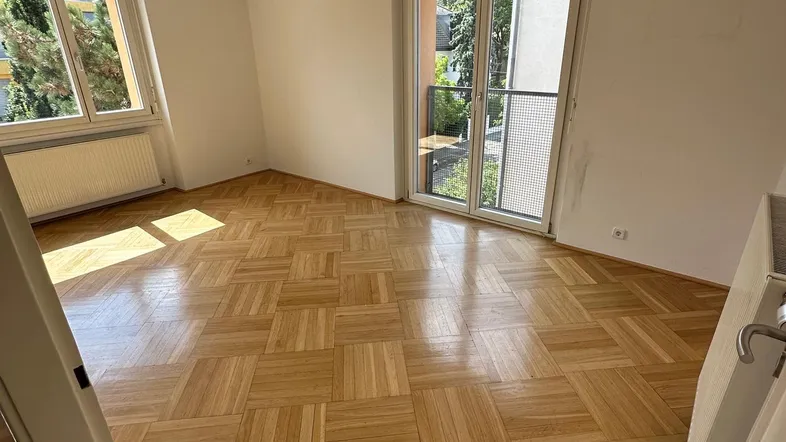 Helles Zimmer mit Fischgrätparkettboden und Zugang zu einem kleinen Balkon.