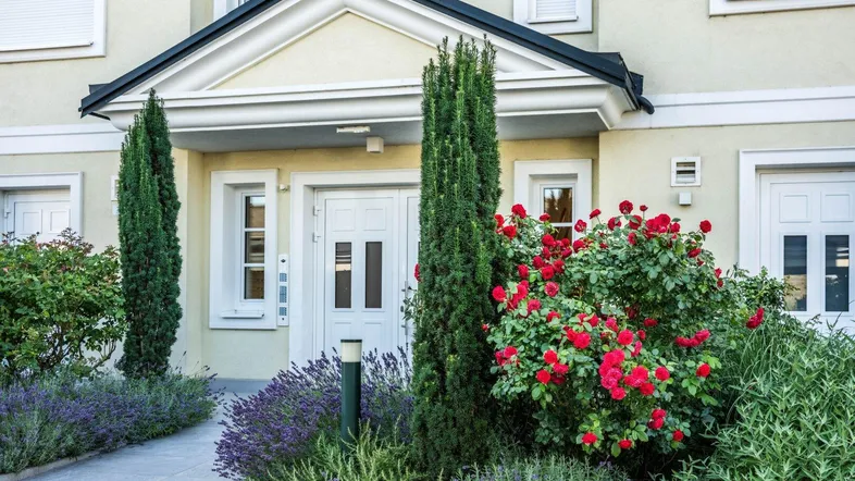 Eingangsbereich eines Gebäudes mit gepflegter Gartenanlage, Rosenbüschen und Zypressen.