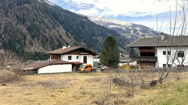 Blick auf Wohnhäuser in einer alpinen Landschaft mit schneebedeckten Bergen im Hintergrund unter bewölktem Himmel.