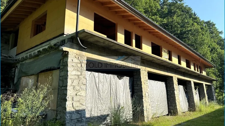 Außenansicht eines im Bau befindlichen Gebäudes mit Steinfundament und Holzkonstruktion, umgeben von Bäumen.