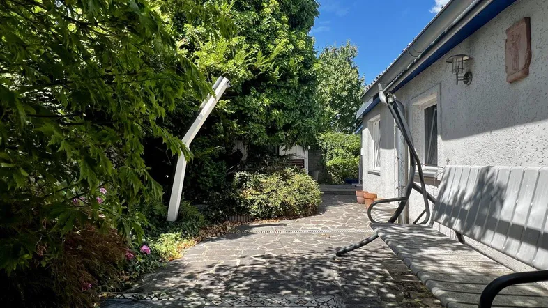 Eine gepflasterte Terrasse mit einer Schaukelbank neben dem Haus, umgeben von viel Grün.