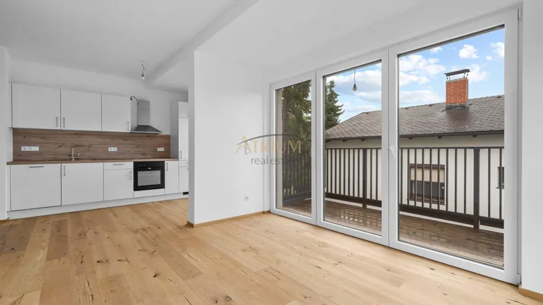 Heller offener Wohnbereich mit integrierter Küche, Holzboden und Zugang zum Balkon.