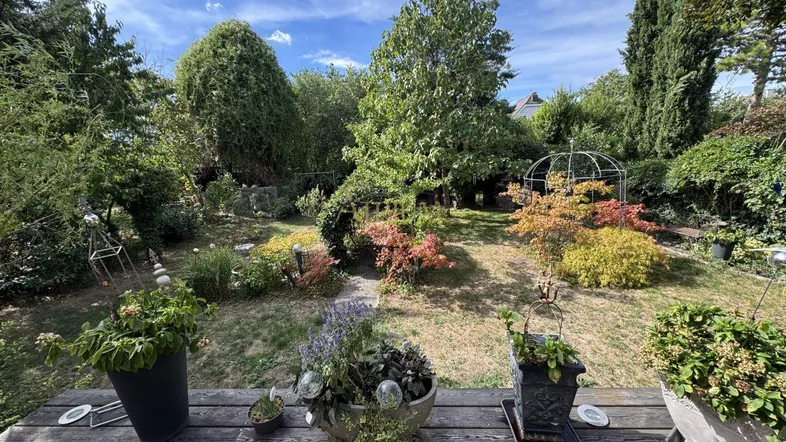 Blick von einer Holzterrasse in einen üppigen, sonnigen Garten mit vielfältiger Bepflanzung und Bäumen.