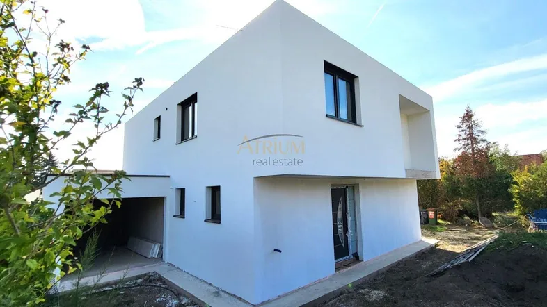 Moderne weiße Hausfassade eines Neubaus mit klaren Linien und dunklen Fenstern, umgeben von Baustelle und Grün.