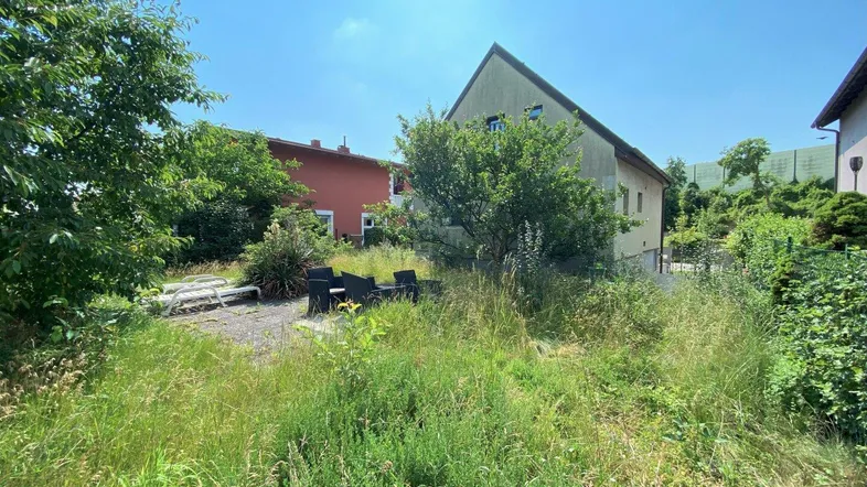 Verwilderter Gartenbereich mit Sitzgelegenheiten und Blick auf die Rückseite des Hauses unter blauem Himmel.
